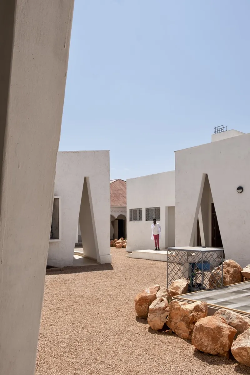 Between buildings: white walls with triangular openings framing the passage, rocks, figure walking through