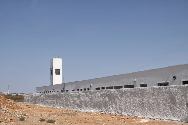 Long facade from distance: hospital behind boundary wall, two wind towers at each end, dry terrain