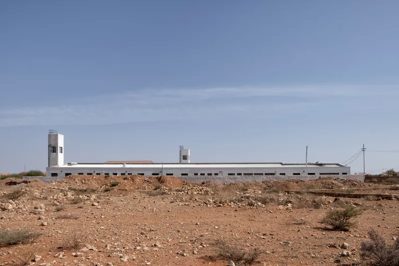 Full hospital from the desert: long white building with mono-pitch corrugated roof, two wind towers, flat horizon