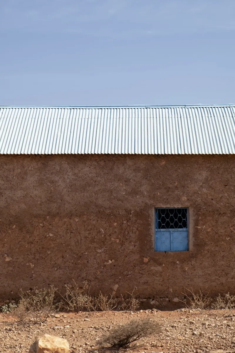 Local vernacular: mud and adobe building with corrugated roof, blue-shuttered window, dry red ground