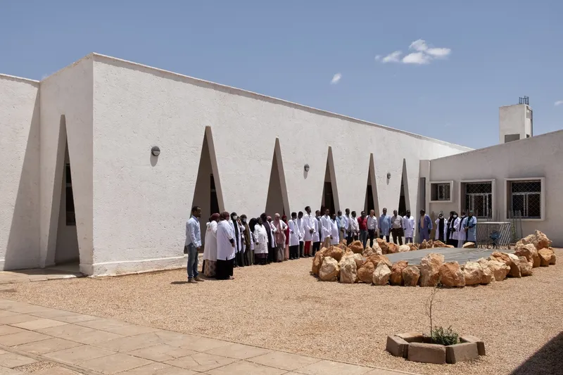 Hospital staff gathered in the courtyard for a group photograph, triangular windows behind, young tree at centre