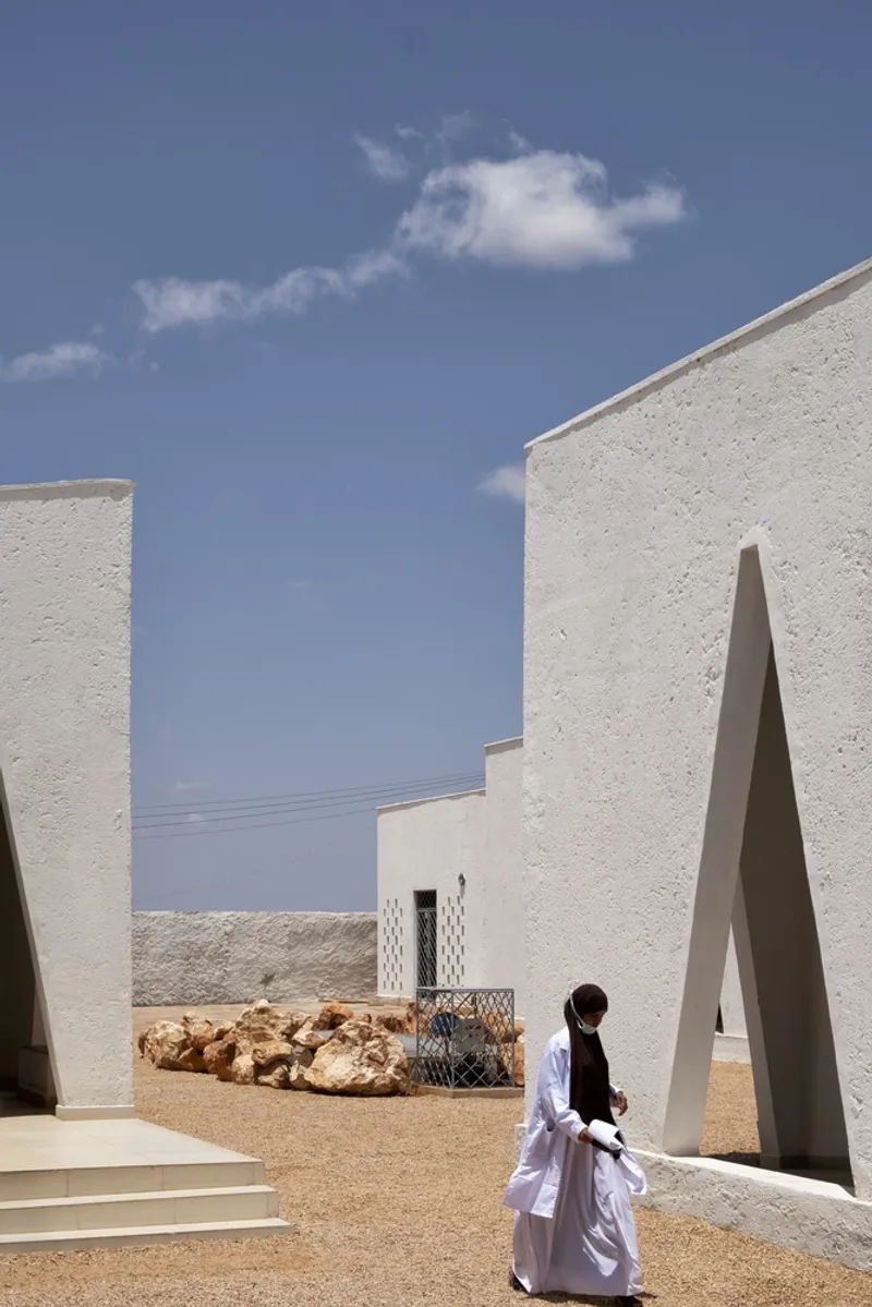 Between buildings: figure walking past triangular openings, white walls, local rocks on the ground