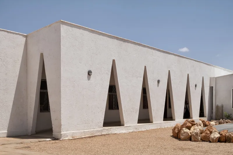 Long facade: row of triangular windows along the courtyard wall, rocks at base, wall-mounted light fixture