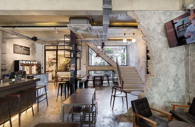 Ground floor bakery: long glass display counter lit from below, exposed concrete walls and ceiling, metal stools, stair to upper floor