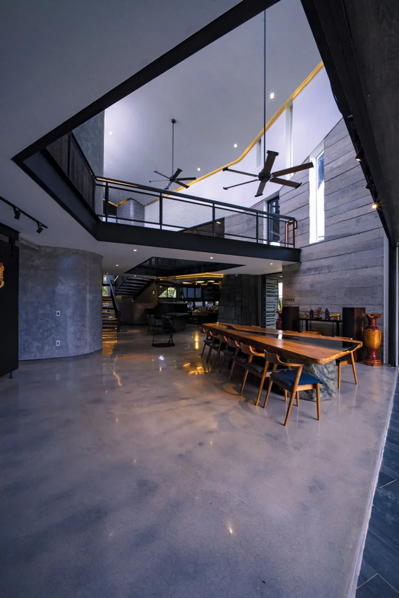 Double-height interior volume with cantilevered steel walkway, concrete walls and dining table at dusk