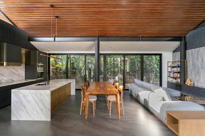 Open-plan living and dining area with timber slat ceiling overlooking a forested courtyard through floor-to-ceiling glazing