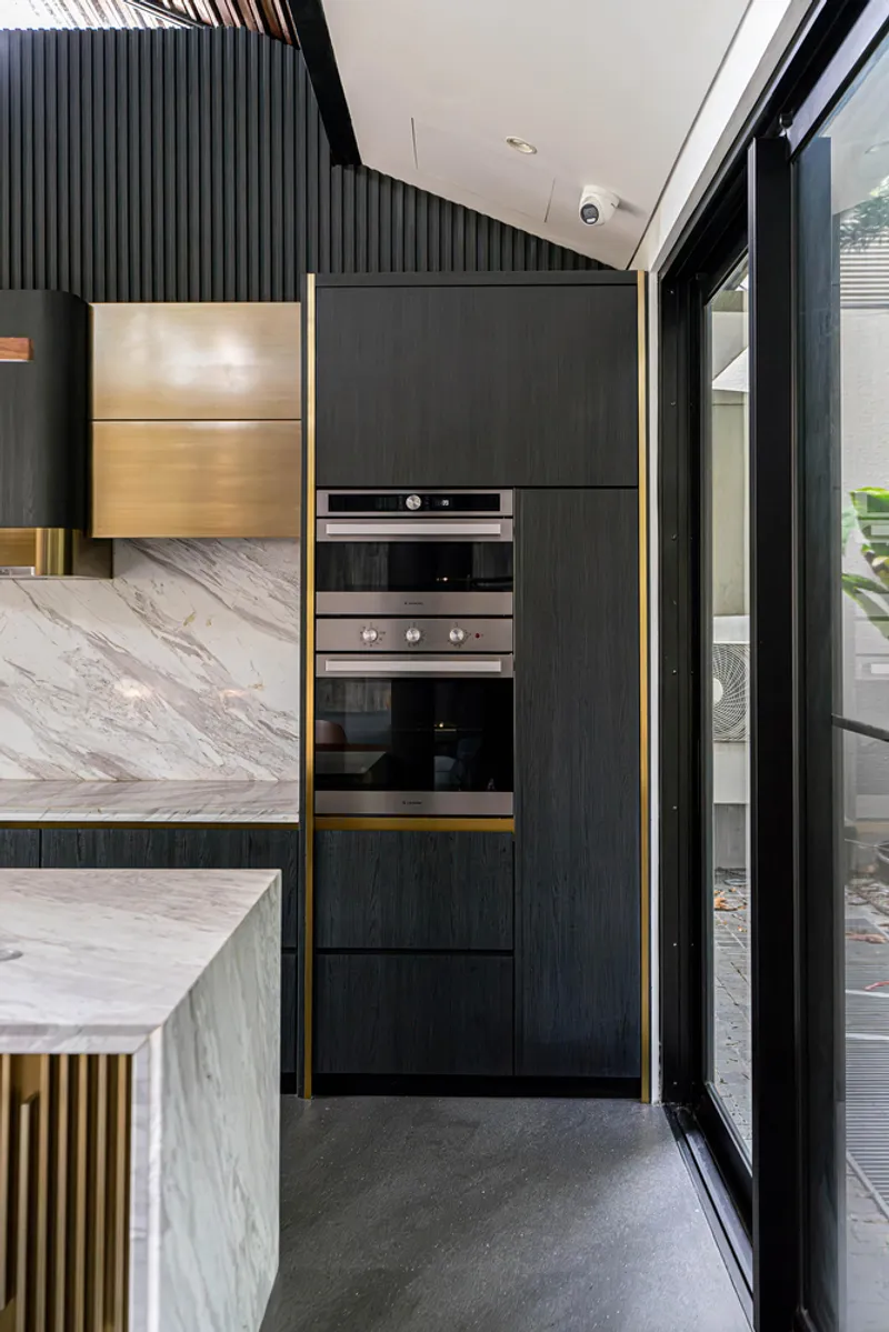 Dark oak cabinetry with brass edge trim housing stacked ovens beside marble backsplash and glass doors