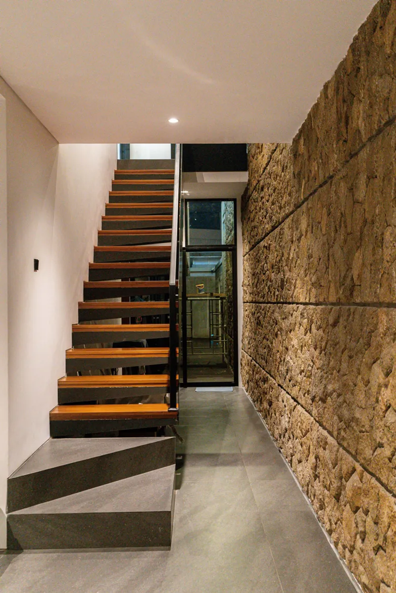 Floating timber staircase along textured stone wall with glass elevator visible beyond the landing