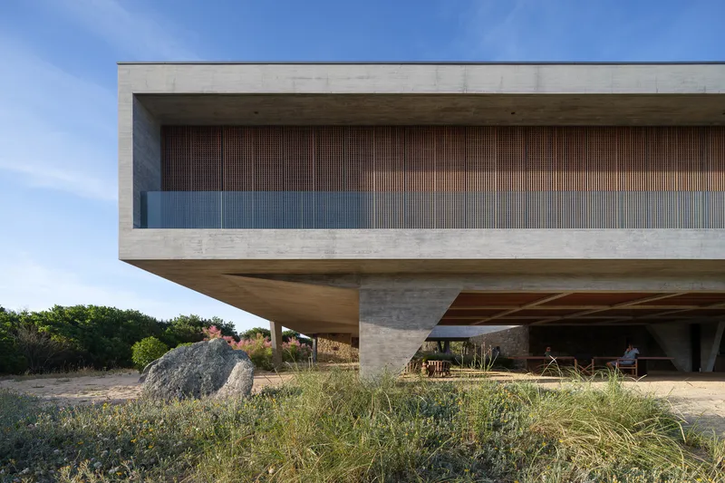 Cantilever detail: exposed concrete soffit and edge beam, timber louvre screen, V-column meeting the ground beside a large boulder