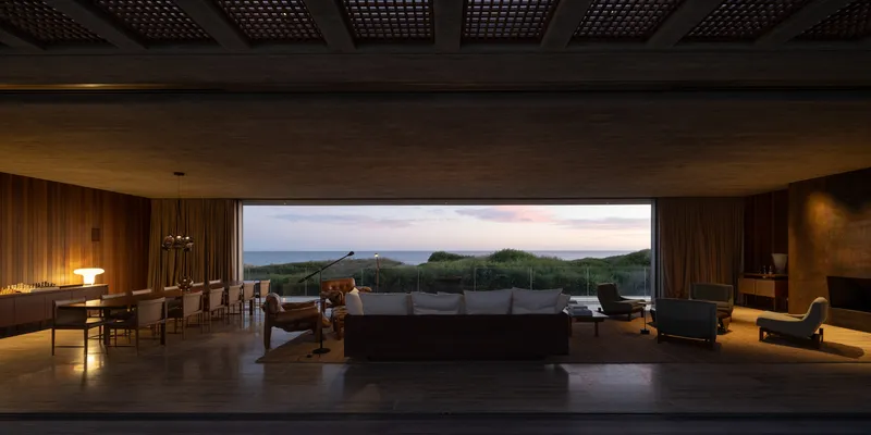 Living room at dusk: timber-lined walls and coffered ceiling, full-width opening framing the dunes and sea, sofa, dining table, fireplace at right