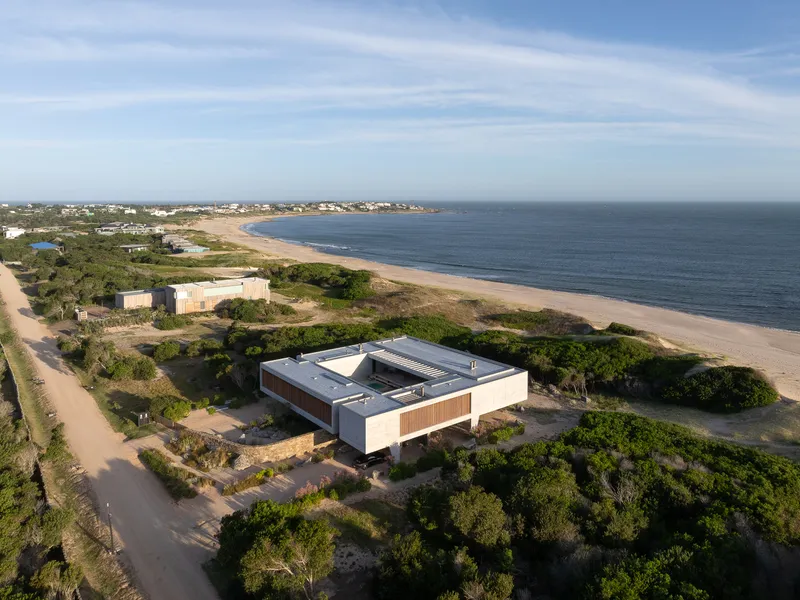 Aerial from the sea: the house sitting between dune cordon and coastal forest, road and neighbouring houses beyond, beach at left