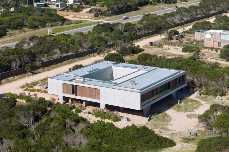 Aerial from the road: flat-roofed concrete volume with central courtyard visible, timber screens, dune vegetation, road and neighbours behind