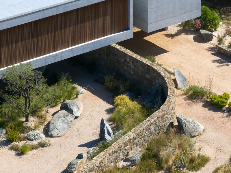 Entry detail from above: curved dry-stone wall guiding the approach, boulders, native shrubs, upper volume cantilevering above