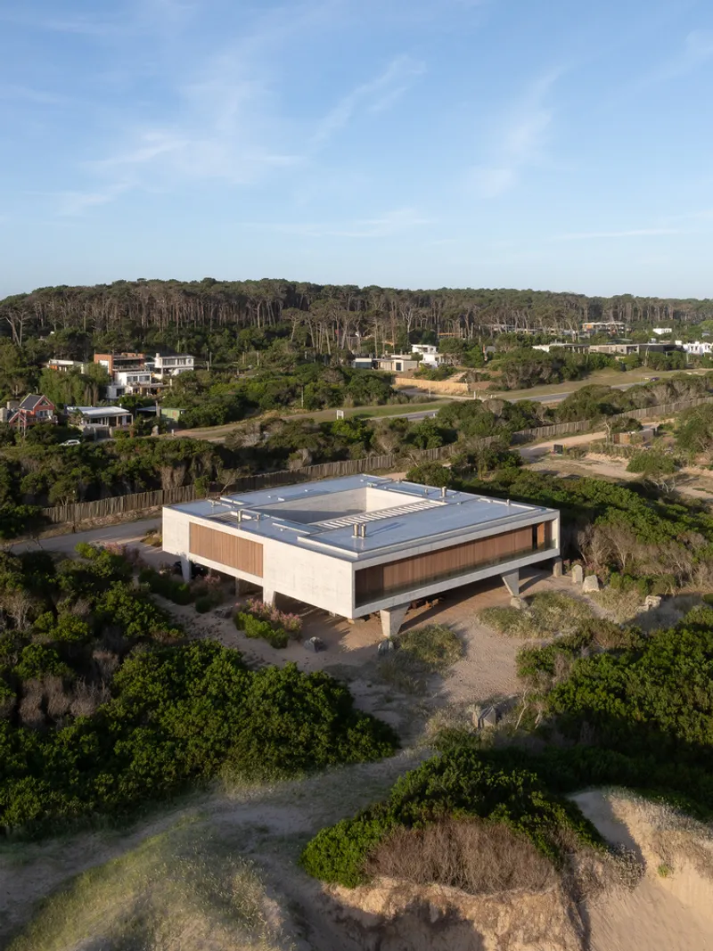 Aerial from the dunes: concrete box with timber screen facing the sea, flat roof, coastal scrub and forest behind, late afternoon light