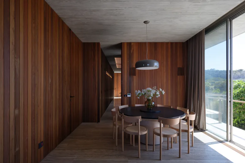 Dining room: timber-panelled walls, exposed concrete ceiling, round table with chairs, pendant light, floor-to-ceiling window to the landscape
