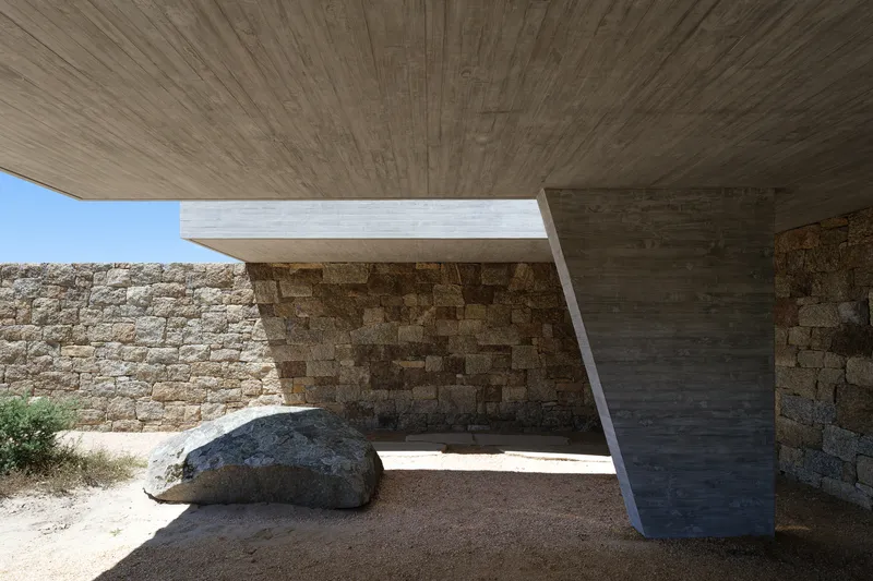 Under the cantilever: exposed concrete soffit, V-column, large boulder left in place, dry-stone wall, sandy ground