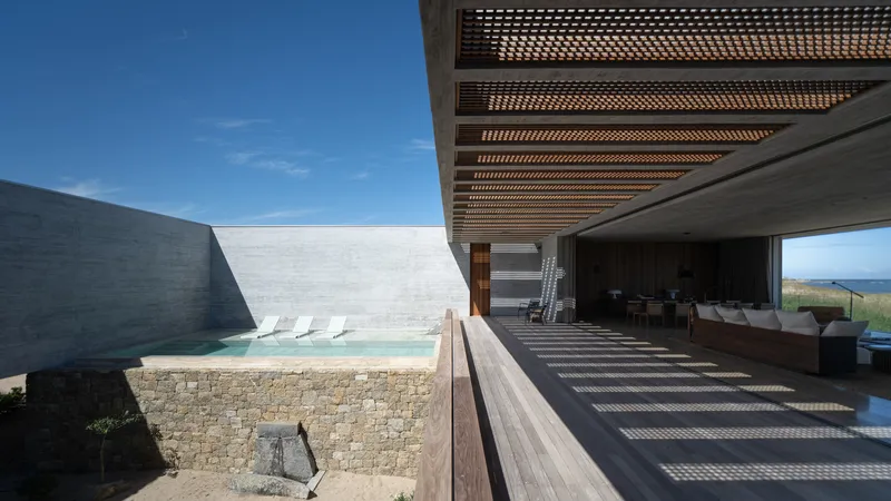 Pool terrace: timber deck, plunge pool with stone wall surround, timber pergola overhead, living room glazing visible at right, dunes beyond