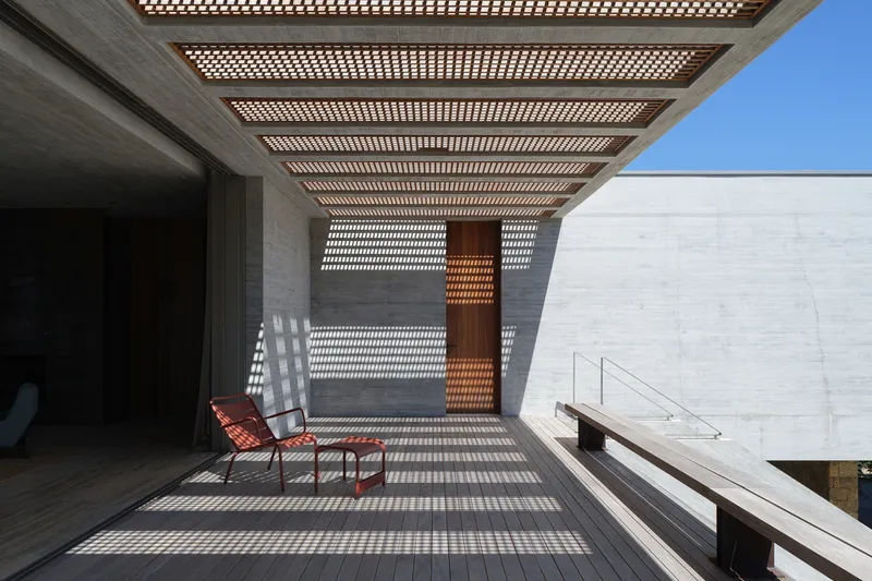 Upper terrace: timber lattice pergola casting striped shadows on the concrete floor, two orange chairs, timber pivot door, concrete walls