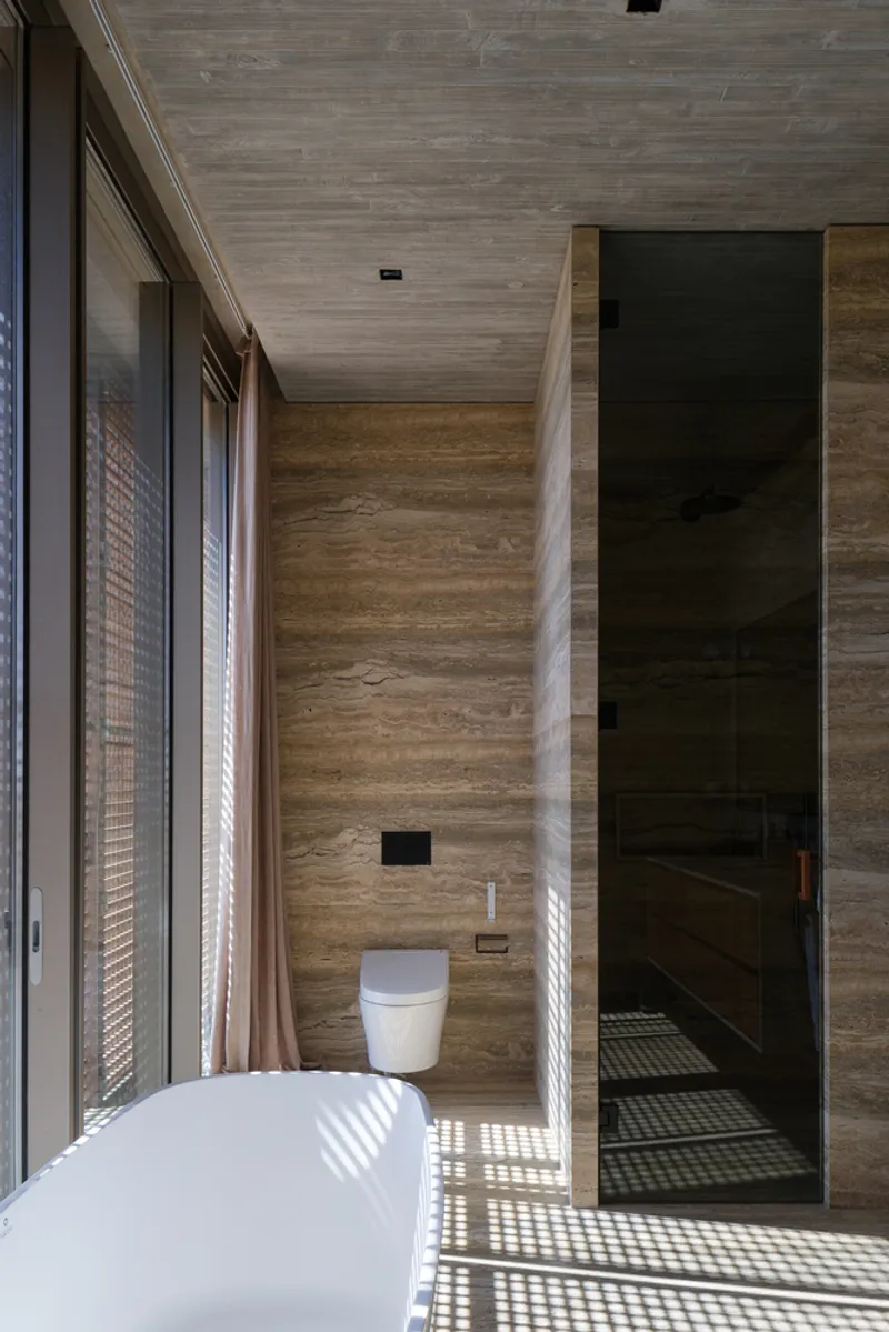 Bathroom: timber-lined walls, exposed concrete ceiling, freestanding white tub, lattice screen casting light patterns, dark stone floor
