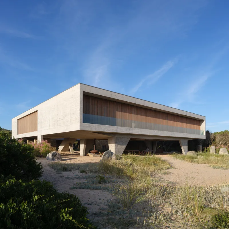 From the south-east dunes: concrete volume with timber screen, boulders in the foreground, coastal scrub, overcast sky