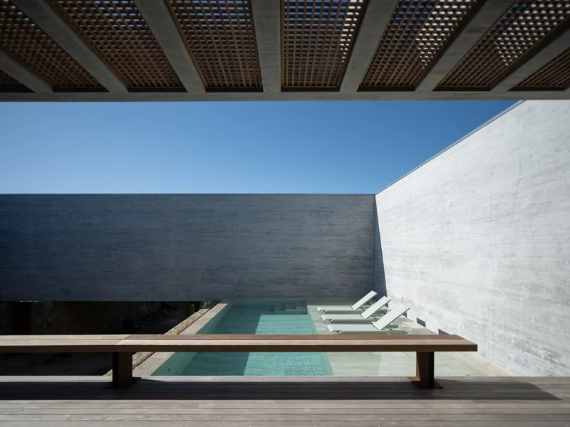 Pool courtyard frontal: turquoise pool, white loungers, timber bench in foreground, high concrete and stone walls enclosing the space, timber pergola above