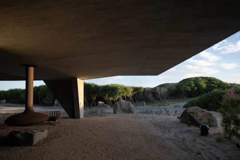 Ground-level terrace: outdoor fireplace column, boulders, sandy ground, dune landscape and scrub visible beneath the concrete soffit