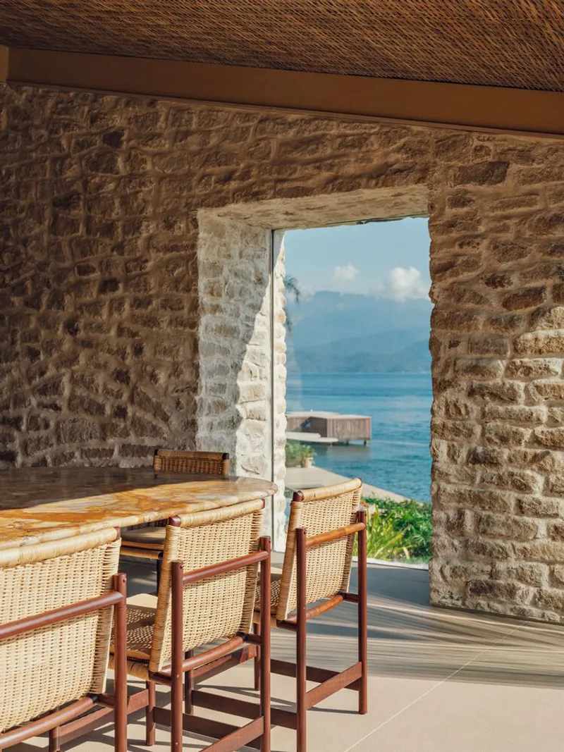 Stone threshold opening framing lake view with distant mountains and timber dock beyond wicker dining chairs
