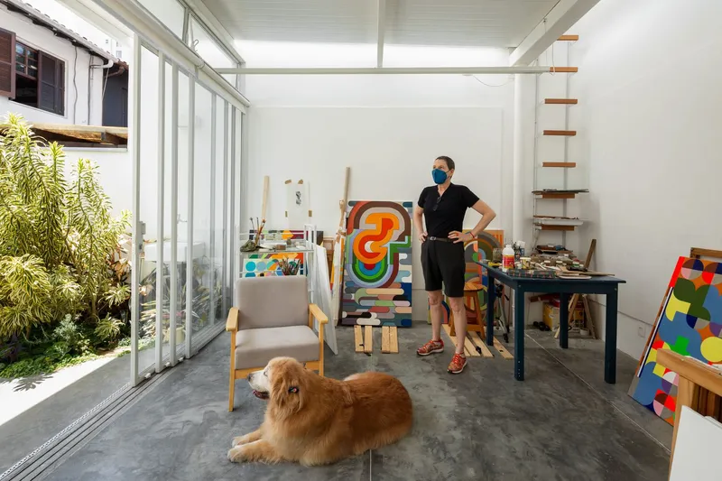 Atelier interior looking out: full-width sliding glass to the garden courtyard, armchairs, easels, paintings