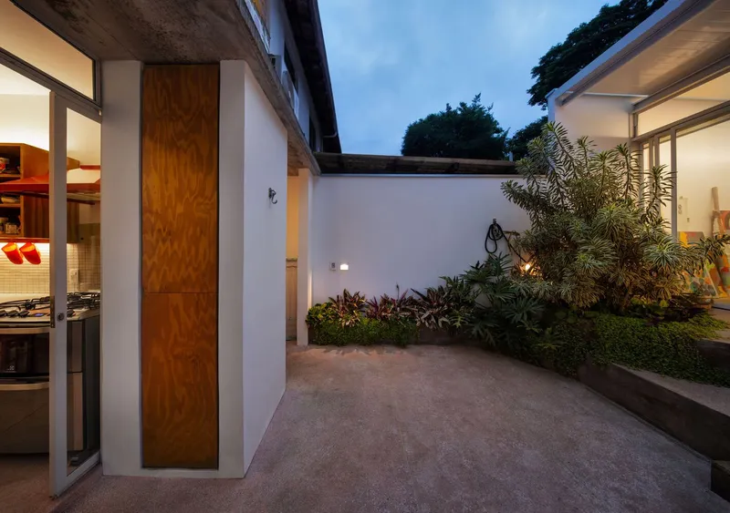 Dusk: courtyard garden lit warmly, plywood pivot door, planting beds, atelier glowing at the back