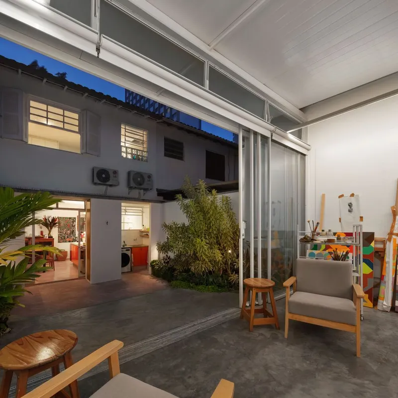 Dusk from the atelier: glass walls open, paintings visible, steel frame and translucent roof lit from inside