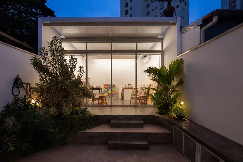 Dusk exterior: the atelier as a glowing glass box at the end of the garden, concrete steps, tropical plants