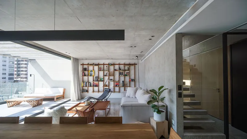 Living room with exposed concrete ceiling, timber bookshelf wall, daybed on terrace and interior staircase ascending