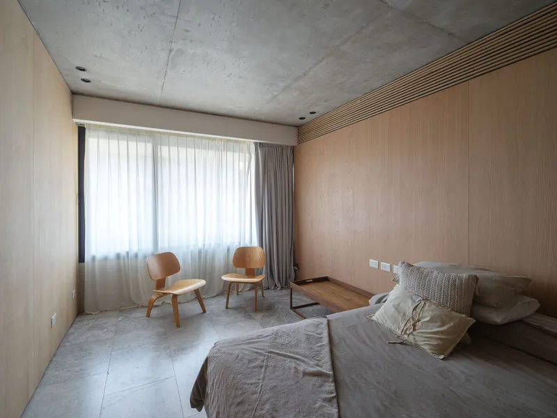 Bedroom with pale timber wall panels, concrete ceiling, sheer curtains filtering daylight, and two plywood lounge chairs