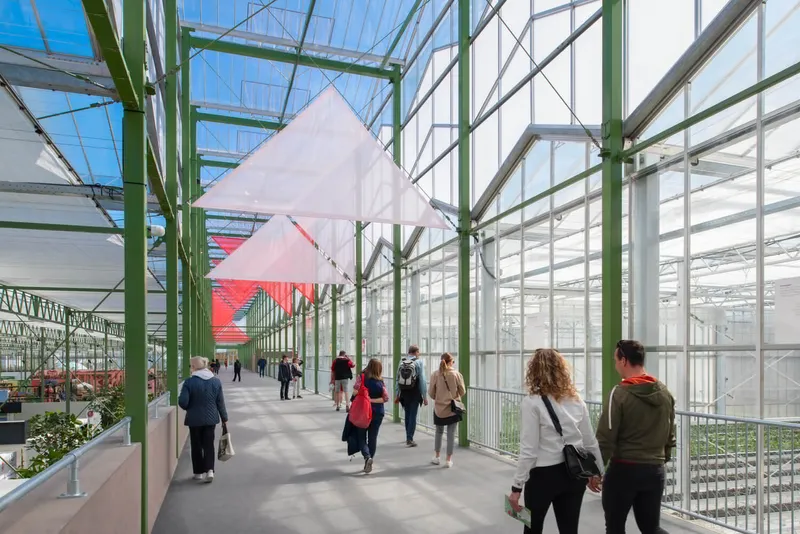 Glazed greenhouse corridor with green steel trusses and visitors walking toward suspended white pavilions