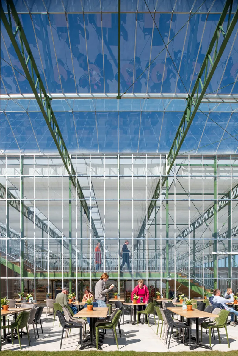 Dining area beneath a transparent roof with green steel structure and visitors at wooden tables in afternoon light