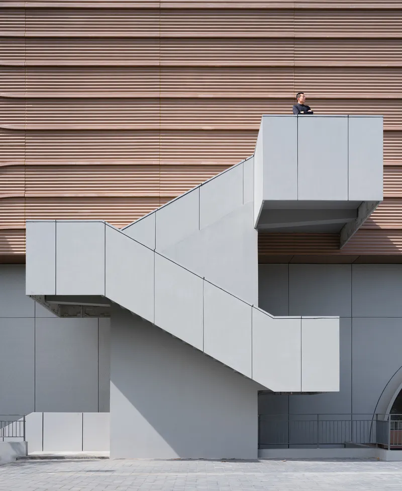 Sports centre front elevation: large white volume with clerestory glazing, landscaped entry, trees, signage