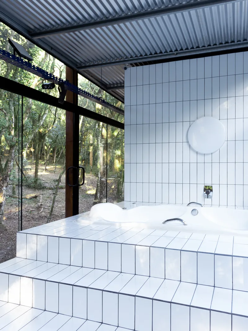 Bath detail: white tile soaking tub with corrugated metal ceiling above, glass corner open to the forest, chrome fixtures