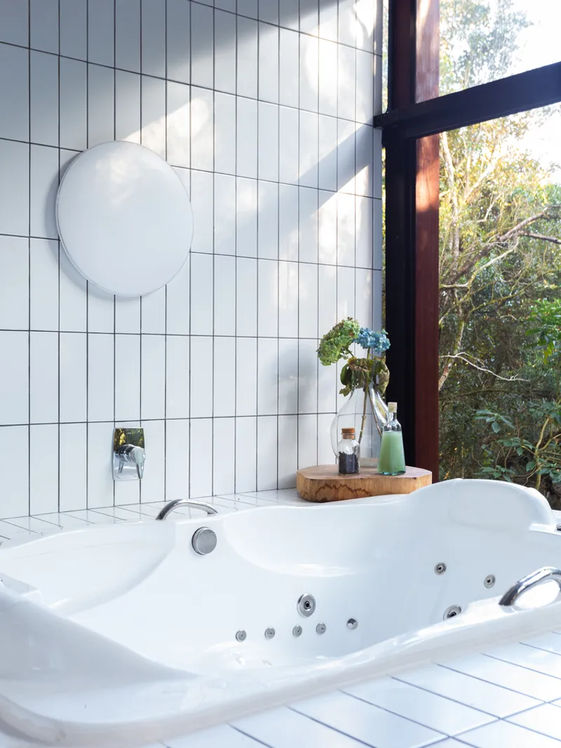 Bath close-up: edge of the white tile tub, round mirror on the wall, timber bath tray, glass wall to the trees, soft daylight