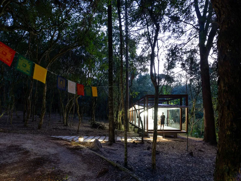 Dusk with prayer flags: coloured Buddhist flags strung between the trees, glass pavilion lit from within, figure standing inside, timber boardwalk, forest