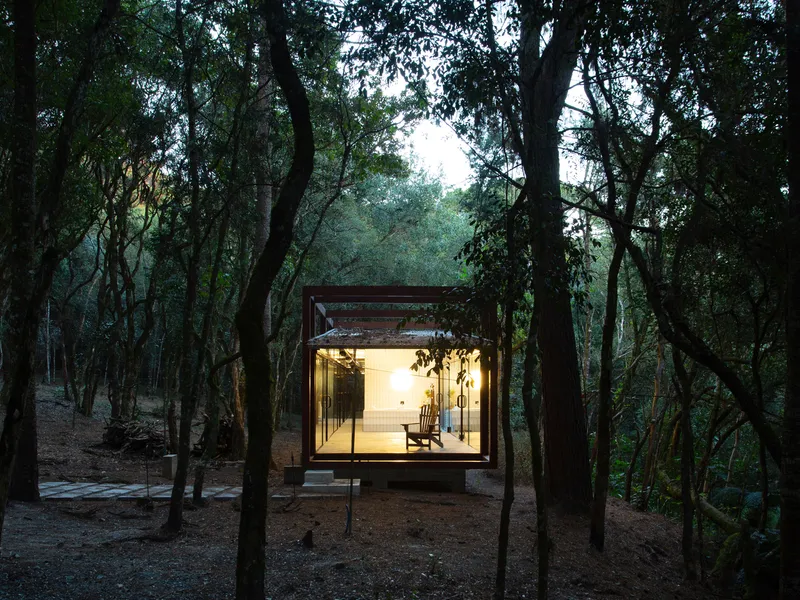 Frontal at dusk: glass pavilion glowing with warm light, timber chair visible inside, corten frame, corrugated roof, dark forest silhouette