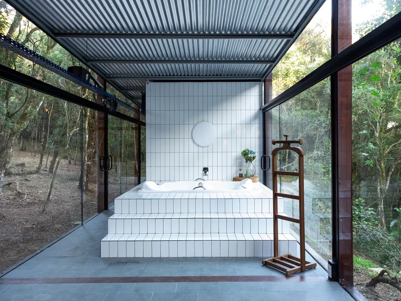 Soaking tub: white tile bath under a corrugated metal roof, full-height glass walls, timber frame, forest visible on all sides, timber towel ladder