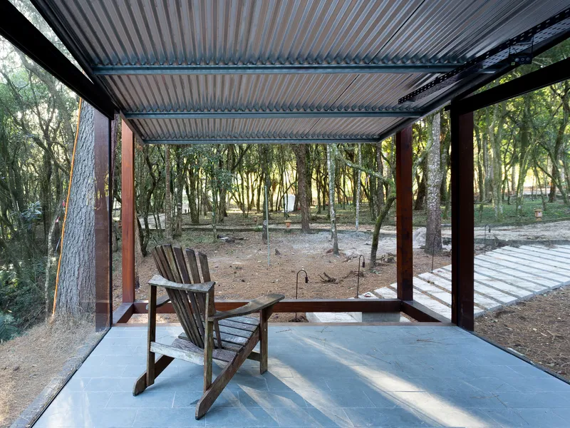 Interior: timber Adirondack chair on stone floor, corrugated metal ceiling, corten steel columns, full-height glass to the forest, warm afternoon light