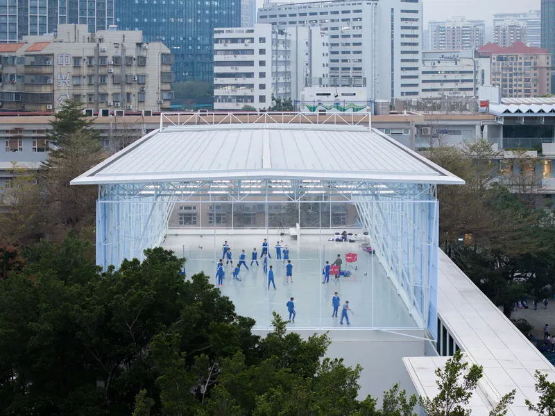 Glass pavilion with performers in blue uniforms gathered on the white floor beneath a standing-seam metal roof