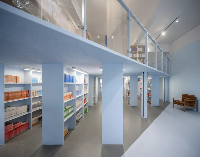 Library interior with pale blue bookshelves arranged in rows beneath a mezzanine with glass railings