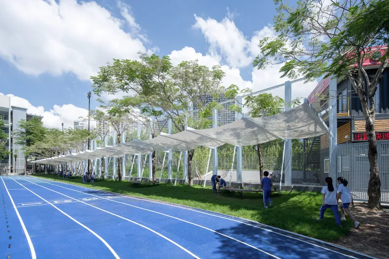 White tensile fabric canopy stretching along a blue running track with trees and students on grass