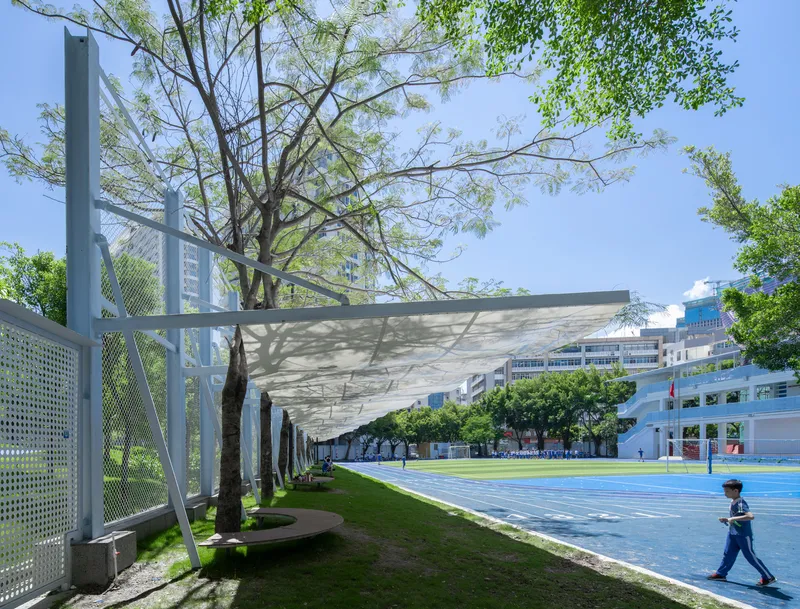 Fabric canopy suspended between white steel columns with tree trunks integrated into the shaded seating area