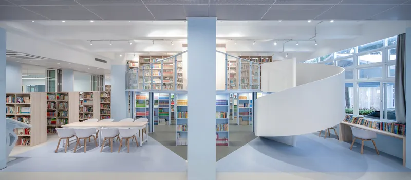 Library reading room with communal table, bookshelves, and a white helical staircase beneath ceiling clerestories