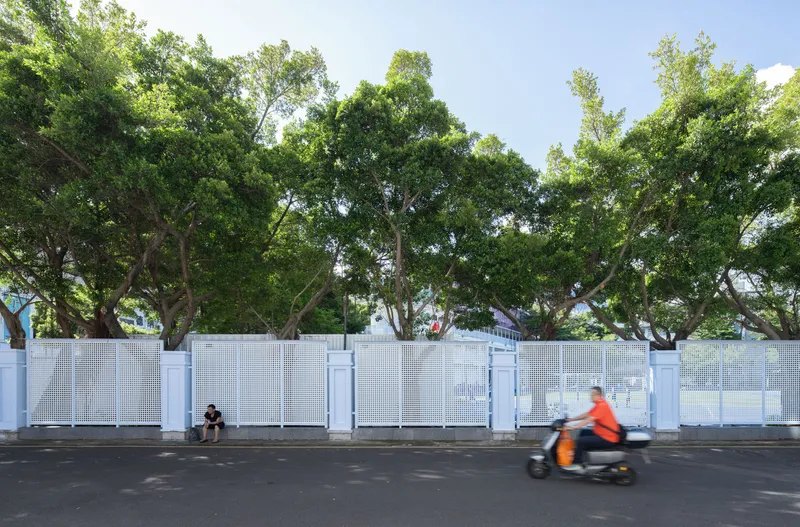 Street view of the white perforated screen wall with a scooter passing and trees overhead