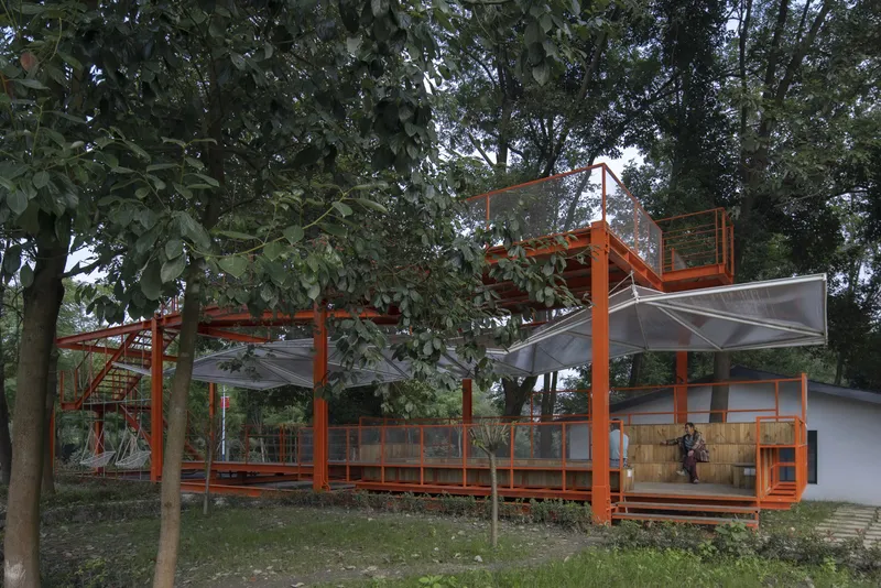 Daytime view: orange steel frame pavilion rising through the tree canopy, timber deck, translucent canopy panels, old mill building behind