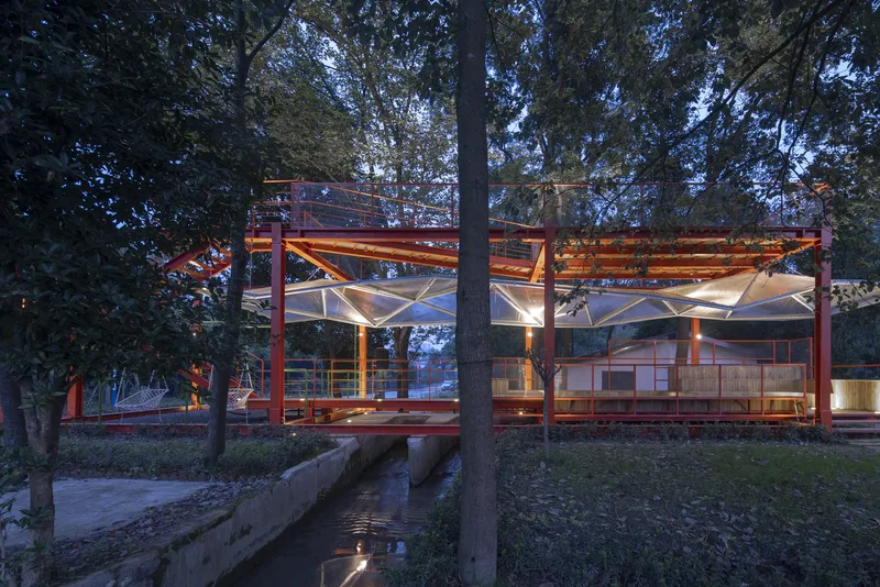 From across the water channel at dusk: orange pavilion lit from within, translucent canopy glowing, trees framing, stone-lined channel in foreground